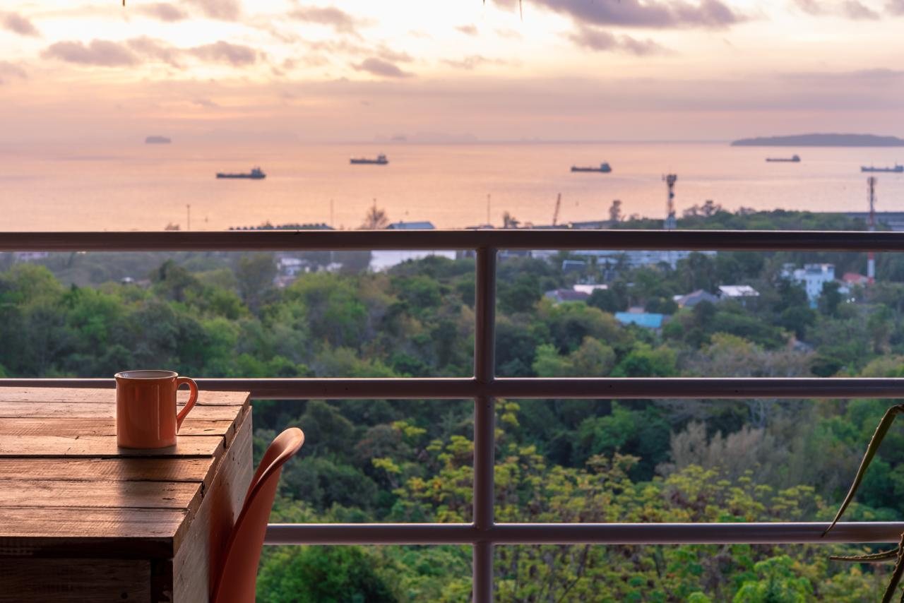 The view from The View balcony: an orange mug on a weathered wooden table, with the Andaman Sea, cargo ships and Cape Panwa rooftops below, all framed by jungle treetops